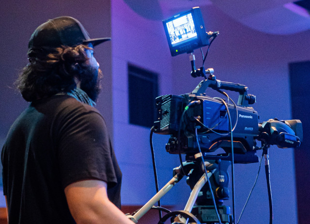 Operador de câmara de boné e barba a filmar é um exemplo de trabalho em televisão.