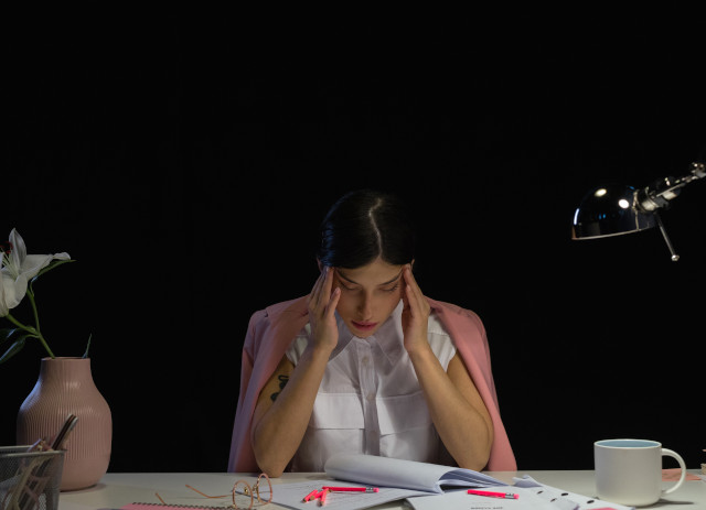 Mulher com camisa branca e casaco rosa nos ombros, segura mãos na cabeça e parece um workaholic.