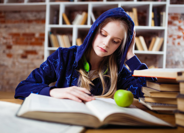 Jovem loura de capuz azul na cabeça a estudar com livro à frente e maçã em cima, talvez a precisar de orientação vocacional.
