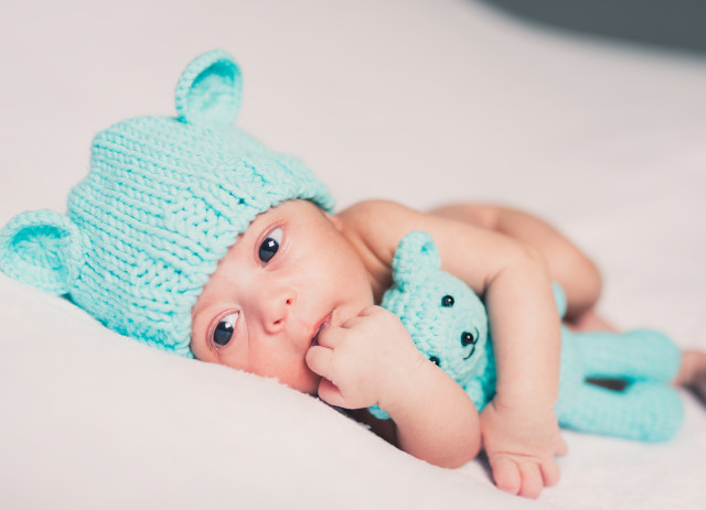 Bebé deitado com mão na boca e gorro e peluche azul-água à espera do Baby Planner.