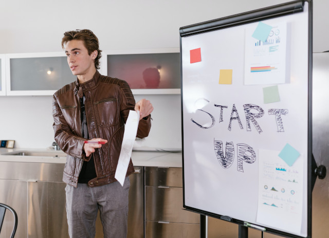 Jovem empreendedor de casaco de cabedal castanho apresenta a sua start-up com um painel e gráficos.