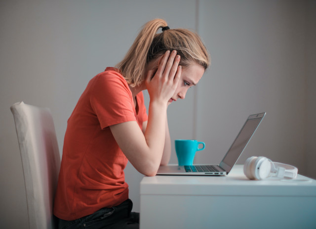 Mulher jovem universitária de t-shirt laranja e a segurar o rosto com as duas mãos, preocupada, debruçada sobre o computador portátil e a ler sobre o complemento de alojamento.