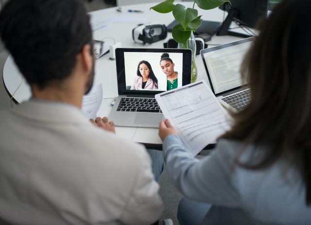 Técnico de Recursos Humanos em frente a um computador, usando óculos e camisola branca, a conversar com colega de camisa azul.