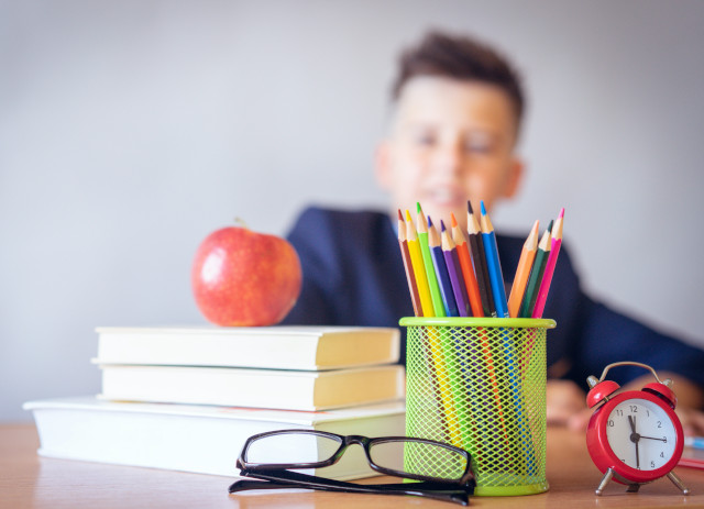 Jovem aluno a olhar para uma secretária com óculos, relógio, canetas, livros e mação em cima a pensar no regresso às aulas.
