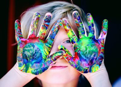 Menino louro de olhos azuis com as mãos pintadas de várias cores à frente do rosto depois das atividades extracurriculares de artes plásticas.