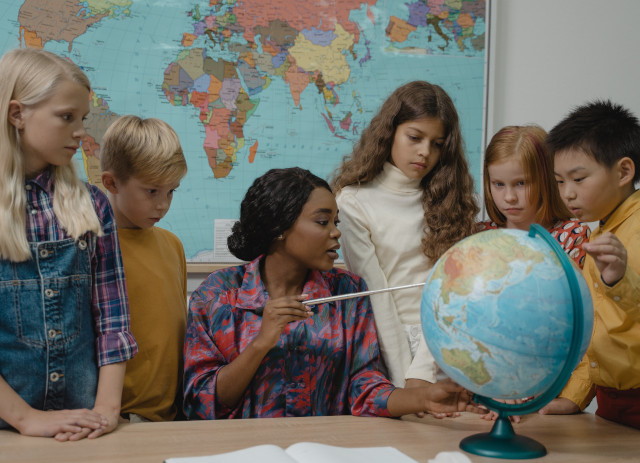 Mulher morena, com camisa colorida e mapa atrás das costas, a mostrar a quatro jovens alunos num globo do mundo quanto ganha um professor em vários países.