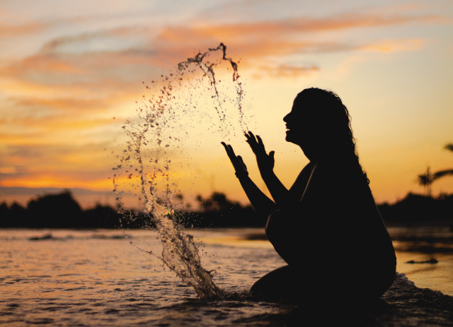 Vulto de mulher sorridente no mar, durante a noite, a atirar água ao ar com as mãos abertas.