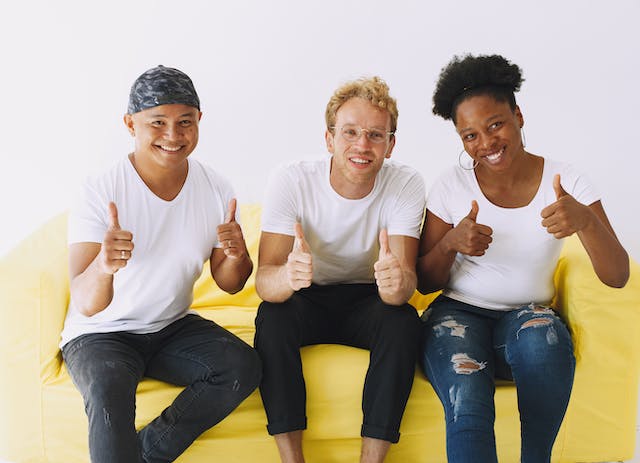 dois homens e uma mulher estão sentados num sofá e dão sinal de aprovação com os dedos
