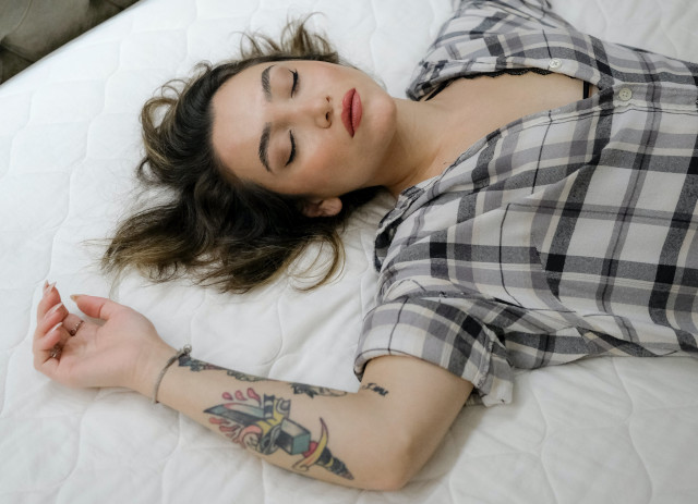 Jovem mulher maquilhada e de olhos fechados, com braço tatuado e pijama preto e branco deitada a testar um colchão.