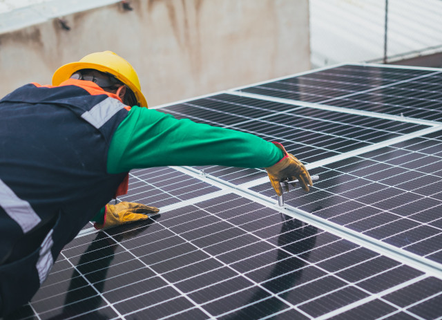 Homem de capacete amarelo e colete azul e verde a instalar painéis solares no âmbito da transição energética.