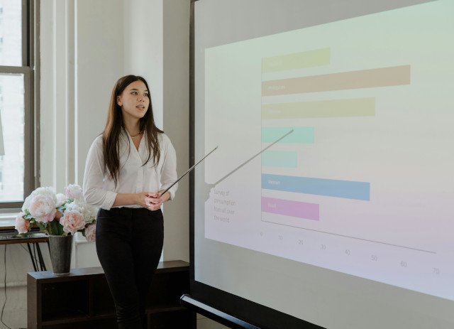 Jovem mulher de ascendência asiática a apontar para painel com tabelas coloridas com dados de cursos de gestão.