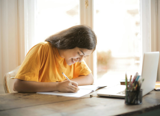 Jovem a escrever numa secretária, com uma caneta e camisola amarela, a sorrir e a apontar dicas de como estudar.