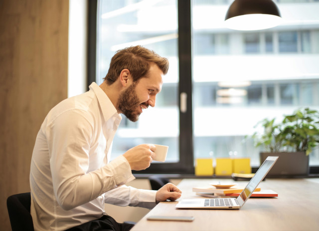 Homem de barba e camisa branca a beber café numa chávena e a sorrir em frente ao computador numa reunião em modelo de trabalho híbrido.