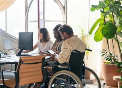 Duas mulheres a mostrarem ofertas de emprego para pessoas com deficiência a um homem numa cadeira de rodas, junto a um computador num escritório com uma planta verde num canto.