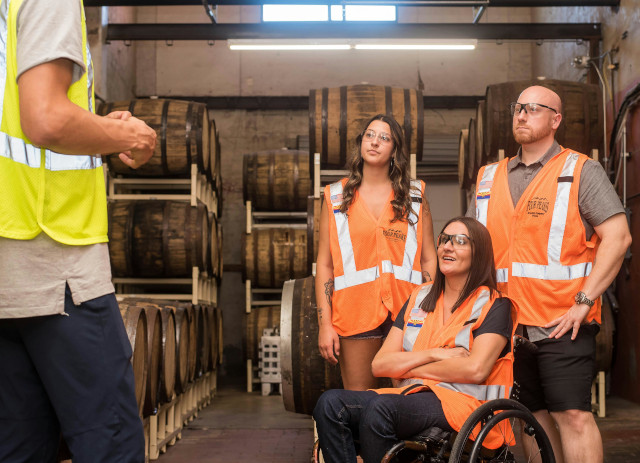 Três trabalhadores com coletes laranja, um dos quais em cadeira de rodas, a ouvir explicações num armazém de alguém de colete amarelo sobre trabalho inclusivo num armazém.