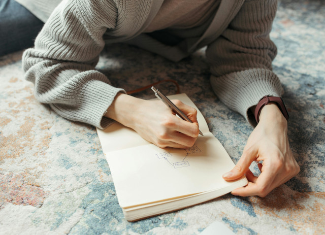 Mulher sentada num tapete com várias cores a escrever num caderno o plano para recomeçar em setembro.