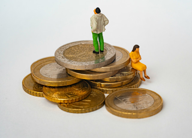 Pilha de moedas de euro com um boneco de um homem de calças verdes e casaco branco no topo e com uma boneca a representar uma mulher mais abaixo, com um vestido amarelo.