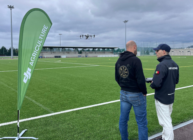 Aluno e formador conversam junto a campo de futebol durante formação de piloto de drones para agricultura.