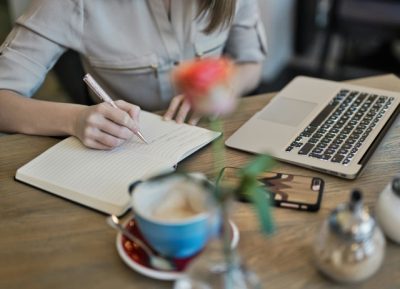 Tronco de mulher a escrever em caderno numa secretária, com um computador à frente e uma flor laranja num vaso em primeiro plano, durante curso de como melhorar a escrita.